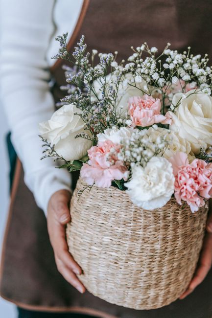 A person holding a round wicker basket filled with a fresh floral arrangement, featuring white roses, pink carnations, baby's breath, and sprigs of lavender, all arranged in a natural, cascading style