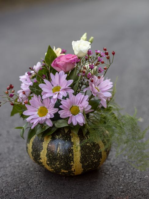 A small, round floral arrangement featuring light pink daisies with yellow centers, white roses, and clusters of tiny pink buds, all set in a black and yellow striped gourd-shaped container. The bouqu