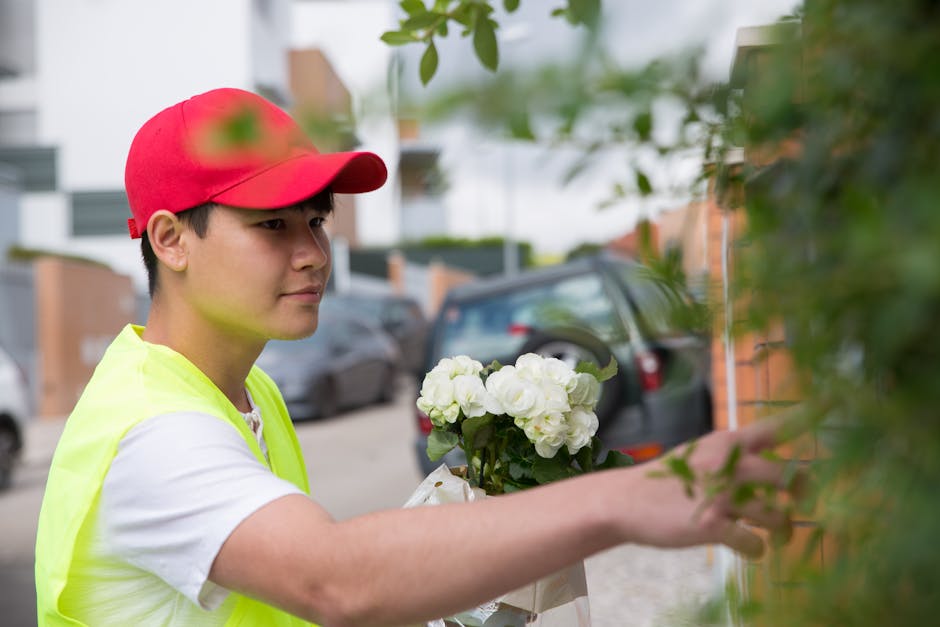 A young delivery person wearing a bright yellow high-visibility vest and a red cap is holding a bouquet of white roses with lush green leaves, preparing to place it against a leafy hedge on a resident
