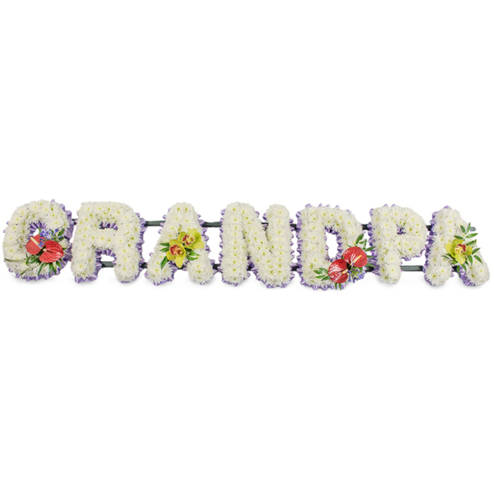The photograph presents a carefully crafted funeral flower tribute spelling out "GRANDPA" in large, bold, three-dimensional letters, aligned in a long row against a stark white background that emphasises their form. Each letter is densely padded with small, white chrysanthemums, packed tightly so they form a plush, cushiony surface. The effect is one of softness and serenity, as though the name itself were gently resting on clouds. The crisp white petals catch the light evenly, giving the tribute a clean and dignified presence that would feel at home in any Mottingham chapel or graveside setting. Tracing the lower edge of each letter is a band of pale lavender ribbon, modest but effective in framing the word and introducing a delicate hint of colour. This soft purple trim adds refinement and helps the individual letters stand out, while a series of dark green support bars beneath and behind the blooms provide the quiet strength needed to keep the piece secure during transit and display. To personalise the name and offer moments of visual focus, four of the letters feature decorative sprays. The "G" and the central "D" host clusters of deep red anthuriums, their glossy, heart-shaped surfaces and yellow spadices emerging from rich green foliage and perhaps a touch of fine filler blooms. These vivid accents symbolise powerful love and remembrance. By contrast, the first and final "A" are adorned with clusters of yellow-green orchids, whose delicate, sculpted petals and intricate throats bring an air of elegance and enduring beauty. This floral lettering would be chosen by a family to honour a much-loved grandfather from Mottingham, a heartfelt blend of tradition, craftsmanship, and quiet emotion expressed entirely through flowers.